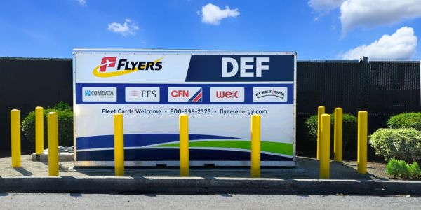 A large white and blue industrial DEF (Diesel Exhaust Fluid) storage tank at a Flyers Energy cardlock station, featuring various fleet card logos and protected by yellow safety bollards.